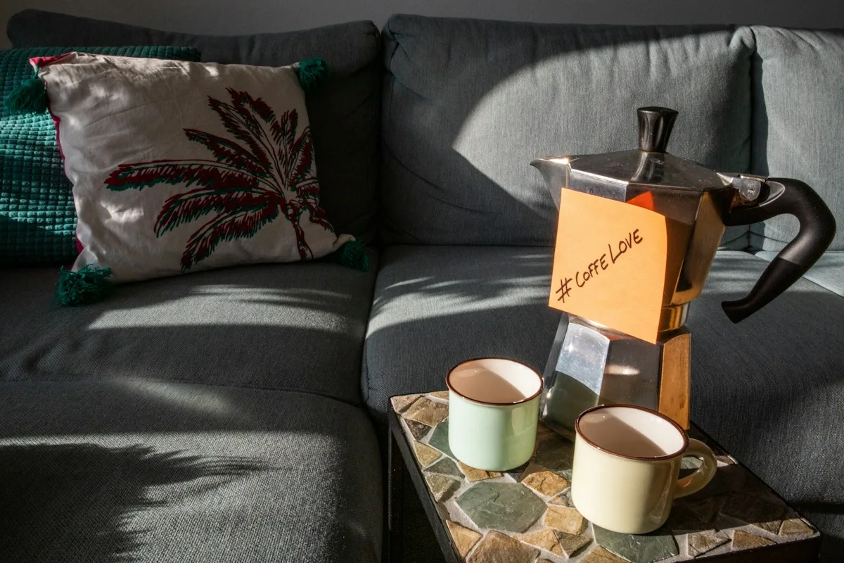 Two cups and a stovetop coffee pot beside a cushioned sofa